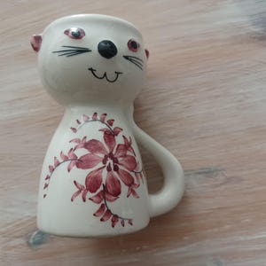 May include: White ceramic egg cup with a cat face and floral design. The cat has black eyes, a black nose, and a black mustache. The flowers are pink and white.