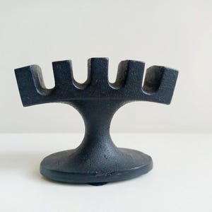 May include: A black, cast iron candle holder with a unique design. The base is round, supporting a stem that leads to a top with five curved slots. The candle holder is approximately 13 cm tall.