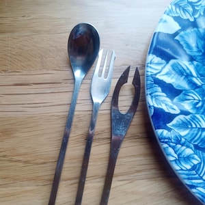 May include: A set of three stainless steel utensils including a small spoon, a two-pronged fork, and a specialised serving tool with an oval cutout, positioned next to a plate featuring a blue and white floral pattern on a wooden surface.