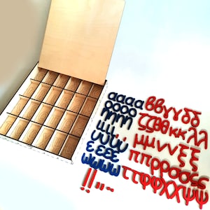 May include: Wooden box with a lid open, revealing compartments for letter tiles. The box is filled with blue and red letter tiles, including the letters a, b, c, d, e, f, g, h, i, j, k, l, m, n, o, p, q, r, s, t, u, v, w, x, y, z. The tiles are arranged in a random order.