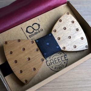 Può includere: Un papillon in legno con pois marroni e un nastro con motivo bianco e nero. Il papillon è in una scatola con le parole "Double O" e "Fatto a mano in Grecia, Creta" stampate su di essa.