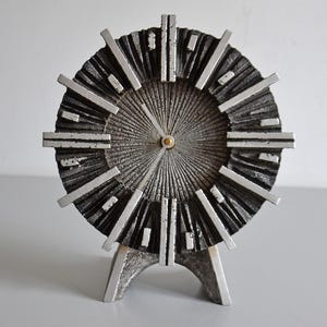 Puede incluir: Un reloj vintage de estilo moderno de mediados de siglo. El reloj presenta una esfera texturizada de color plateado con líneas radiales y marcadores de hora rectangulares. Se apoya en una base plateada de tres patas. Las manecillas son doradas.