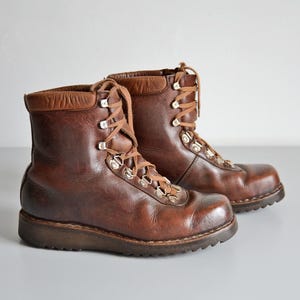 May include: A pair of brown leather ankle boots featuring a smooth finish, a padded collar, and a lace-up closure with silver-toned metal eyelets and hooks. The boots have a thick, dark brown rubber sole with a textured tread pattern for grip.