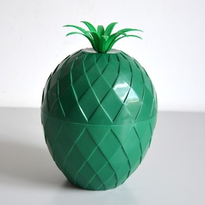 Puede incluir: Un recipiente de plástico verde con forma de piña que presenta un patrón de diamantes texturizado en el cuerpo y una tapa extraíble rematada con una pequeña corona de hojas verdes puntiagudas. El objeto tiene un acabado liso y brillante y forma redondeada.