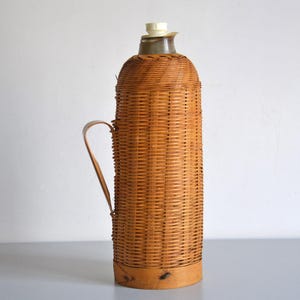 Könnte beinhalten: Eine geflochtene Weide-Thermosflasche in hellbrauner Farbe mit einem Griff. Die Flasche hat einen weißen Plastikdeckel.