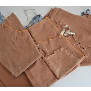 May include: Several brown fabric aprons with blue and white ties. The aprons are made from a robust material and have a simple design. The edges of the aprons are frayed, giving them a rustic appearance.