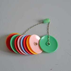 May include: A set of colourful, circular plastic sink stoppers in red, green, pink, blue, yellow, and orange. The stoppers are linked by a silver chain and a green plastic bead.