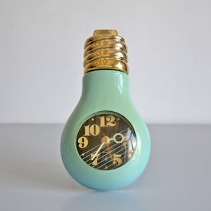 May include: A light mint green lightbulb-shaped clock with a metallic gold screw base. The clock face is circular, featuring a dark background with large gold numerals, thin white diagonal lines, and gold-toned hour, minute, and second hands.