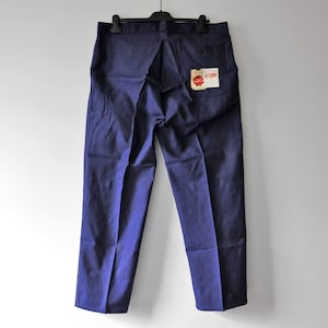 May include: A pair of solid navy blue trousers hanging on a black hanger. The pants feature a straight-leg cut, a waistband with belt loops, and a single back pocket on the right side. A small white rectangular tag is attached to the back pocket.