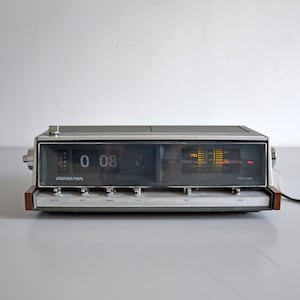 Puede incluir: Un radio reloj de solapas Panasonic rectangular con parte superior gris oscuro, panel frontal metálico plateado y detalles laterales en tono madera. La pantalla muestra la hora 0:08 en un mecanismo de solapas junto a un dial de radio analógico amarillo.