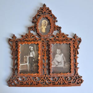 Pode incluir: Uma moldura de madeira castanha para várias fotografias com detalhes intrincados de volutas e filigrana. A moldura contém três fotografias a preto e branco separadas: um retrato oval no topo e dois retratos retangulares abaixo, com bordas recortadas.