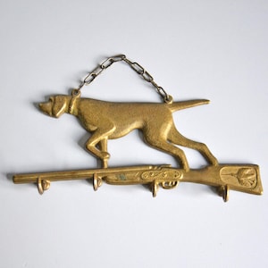 May include: A vintage brass key holder featuring a dog silhouette above a rifle-shaped base with three hooks. The dog is connected to a chain for hanging. The key holder is a decorative item.