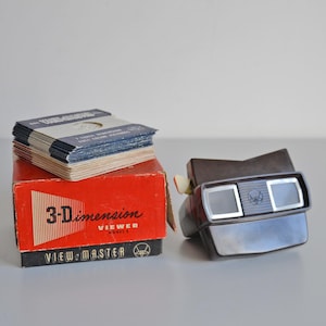 Op de afbeelding: Vintage View-Master Model E met een stapel 3D-beeldrollen en een rode doos. De View-Master is bruin en zilverkleurig met twee oculairen. Op de rode doos staat "3-Dimension Viewer".