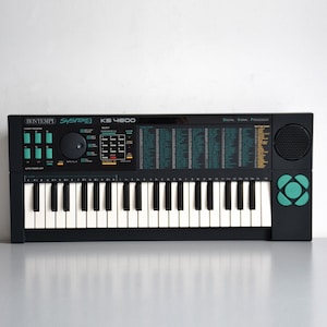 以下が含まれることがあります： 白と黒の鍵盤を備えた黒いBontempi System 5 KS 4600デジタルキーボード。コントロールパネルには、ティール色のボタン、円形のダイヤル、テンキー、楽器音のリストが印刷されています。右側には円形のスピーカーとティール色のロゴがあります。