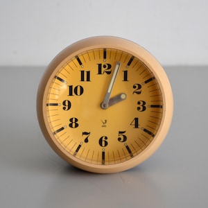 May include: A round analogue alarm clock with a light beige casing and a yellow face. The clock features black Arabic numerals from 1 to 12, black minute markers, and two silver-toned hands. A small bird logo is printed above the number 6.