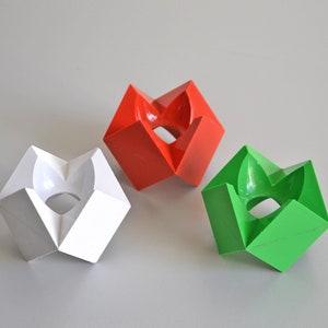 May include: Three geometric candle holders in white, red, and green. Each holder has a unique, angular design with a central circular opening. The holders are arranged on a light gray surface, showcasing their vibrant colors and modern aesthetic.