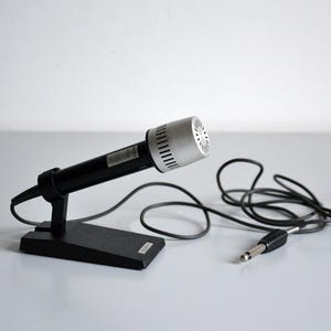 Puede incluir: Un micrófono vintage con cabezal plateado y cuerpo negro, montado en un soporte negro. El micrófono está conectado a un cable negro con un conector jack de 6,35 mm. El diseño es sencillo y funcional.
