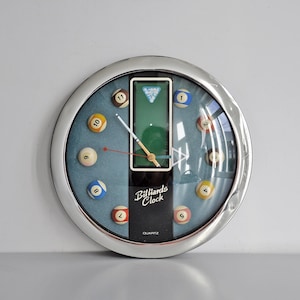 Puede incluir: Un reloj de pared redondo con marco de cromo pulido y esfera azul que presenta bolas de billar en miniatura como marcadores de hora. Un panel vertical negro central muestra el texto Billiards Clock y Quartz, con un gráfico de mesa de billar.