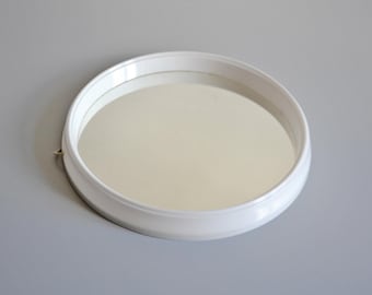 May include: A round, white-framed mirror with a reflective surface. The mirror is set against a light gray background, creating a clean and minimalist aesthetic. The frame has a simple, rounded design.