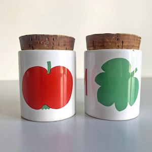 May include: Two white ceramic jars with cork lids. The jar on the left has a red apple design. The jar on the right has a green leaf design.