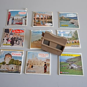 Op de afbeelding: Een verzameling vintage View-Master-schijven met reisbestemmingen zoals Denemarken, Frankrijk en Vaticaanstad. Een bruine View-Master-viewer is ook aanwezig. De schijven tonen kleurrijke afbeeldingen van bezienswaardigheden en landschappen.