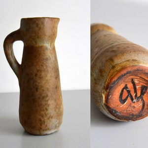 Puede incluir: Una pequeña jarra de cerámica marrón con un esmalte moteado. La jarra tiene un asa y un cuerpo redondeado. La base de la jarra está firmada con "Alf".