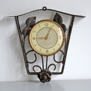 May include: A vintage wall clock with a white face and black Roman numeral markings. The clock is surrounded by a decorative wrought iron frame with a floral design.
