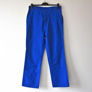 Può includere: Pantaloni blu brillante appesi a una gruccia nera contro un muro bianco. I pantaloni sono di colore pieno, con un classico design a cinque tasche. Il tessuto sembra essere cotone robusto o materiale simile, adatto all'abbigliamento casual.
