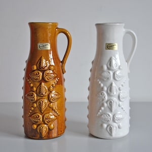 May include: Two ceramic vases, one amber and one white, with handles and embossed leaf designs. The amber vase has a glossy finish, while the white vase has a matte finish. Both have a Europa sticker.