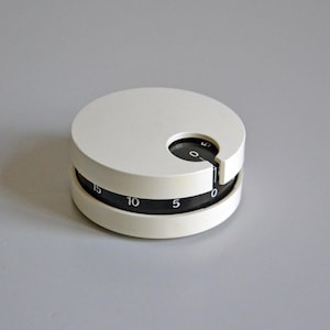 Peut inclure: Un minuteur de cuisine blanc et circulaire avec un cadran rotatif noir. Le cadran comporte des chiffres blancs de 0 à 15. Le minuteur a une petite découpe pour faire tourner le cadran. Le minuteur est sur une surface grise.
