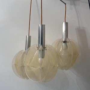 Puede incluir: Tres lámparas colgantes con pantallas esféricas de color crema. Cada lámpara tiene una parte superior cilíndrica plateada y un cable marrón. Las pantallas tienen un diseño geométrico. Las lámparas cuelgan contra una pared blanca.