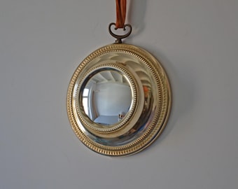May include: A round, silver-colored mirror with a decorative gold-colored rim and a brown leather strap for hanging. The mirror reflects the room. The mirror is a decorative wall hanging.