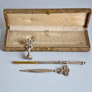 Op de afbeelding: Een antieke schrijftafelset met een zilverkleurige briefopener, pen en zegel, gepresenteerd in een open, verouderde doos. De set heeft bloemmotieven en een goudkleurige penpunt.