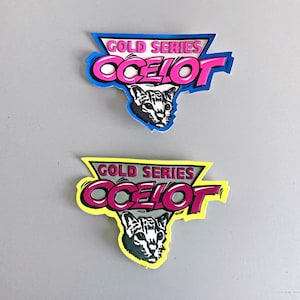 Peut inclure: Deux autocollants avec le texte "GOLD SERIES OCELOT" en rose et blanc, avec un graphique de tête d'ocelot noir et blanc. Un autocollant a un contour bleu, l'autre a un contour jaune.