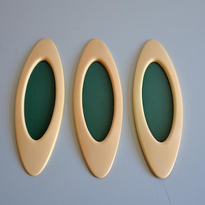 May include: Three oval-shaped picture frames with a cream-colored border and a dark green interior. The frames are arranged in a row against a light gray background. The frames have a smooth, glossy finish.