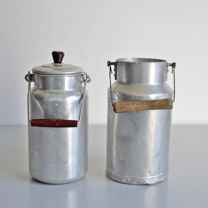 Pode incluir: Duas latas de leite vintage de alumínio. Uma tem tampa com botão marrom escuro e alça marrom escuro. A outra lata está aberta com alça de madeira. Ambas têm alças de metal.