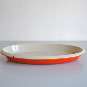 May include: A round, shallow ceramic baking dish. The exterior is bright orange, the interior cream-coloured, and the rim white. The dish is empty and sits on a white surface. Suitable for baking pies and quiches.