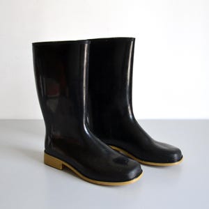 Puede incluir: Un par de botas de goma negras con un acabado brillante. Las botas son de media caña y tienen la puntera cuadrada. Las suelas son de color beige claro. Las botas están sobre una superficie gris claro.