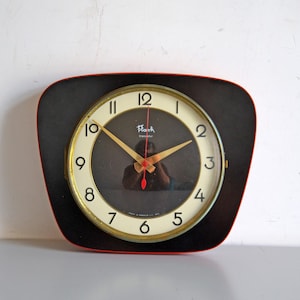 May include: A wall clock with a black, rounded-square frame and a red outer edge. The clock face features a cream-coloured circular dial with black Arabic numerals, gold-toned hands, a red second hand, and the text Flash Transistor.
