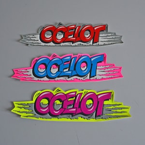Könnte beinhalten: Drei Aufkleber mit dem Wort "OCELOT" in einer stilisierten Schrift. Die Aufkleber befinden sich auf einem grauen Hintergrund. Der obere Aufkleber ist rot, der mittlere Aufkleber ist blau und der untere Aufkleber ist pink.