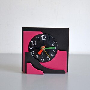 May include: A square desk clock featuring a black and hot pink layered geometric frame. The circular clock face is black with white numerals from 1 to 12. It includes a green hour hand, a red minute hand, and a yellow wavy second hand.