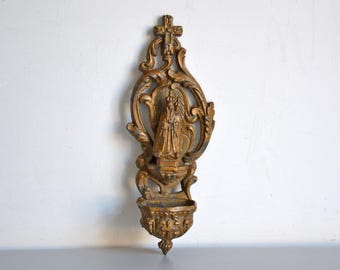 May include: A gold-colored metal wall-mounted holy water font with an ornate design featuring a figure of the Virgin Mary. The font has a small basin for holding holy water.