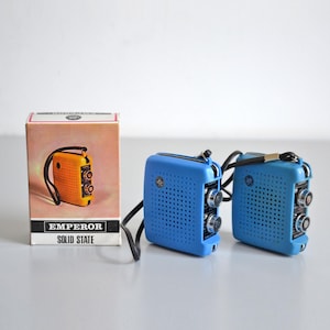 Könnte beinhalten: Zwei tragbare blaue Transistorradios mit schwarzen Handschlaufen, ausgestattet mit einem perforierten Lautsprechergitter und zwei silbernen Drehknöpfen an der Seite. Daneben steht ein Karton mit der Aufschrift EMPEROR SOLID STATE, der ein orangefarbenes Radio zeigt.