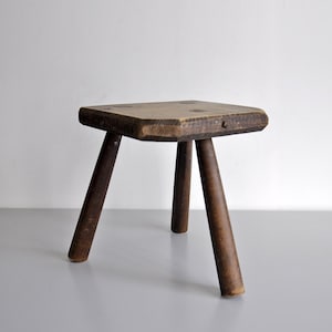 Puede incluir: Un taburete pequeño de madera de tres patas con un acabado marrón oscuro. El taburete cuenta con un asiento grueso y rectangular con esquinas redondeadas y tres patas cilíndricas robustas que se inclinan hacia afuera para dar estabilidad.