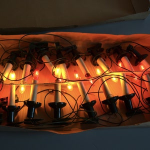 Può includere: Un set di luci natalizie vintage a forma di candela in una scatola di cartone. Le luci sono collegate tra loro da un filo nero e hanno lampadine arancioni. Le luci sono in un groviglio nella scatola.