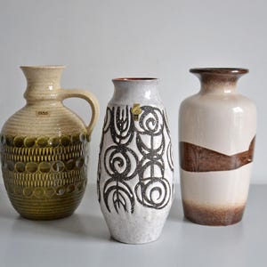 Peut inclure: Trois vases en céramique. Le vase de gauche est beige avec une anse et des détails vert olive. Le vase du milieu est blanc avec un motif tourbillonnant noir. Le vase de droite est beige avec un accent marron.