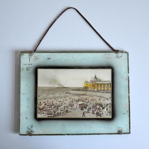 Może przedstawiać: Vintage oprawiony wydruk przedstawiający scenę nadmorską z molo i plażowiczami. Wydruk umieszczony jest w prostokątnej ramie z postarzanym, jasnoniebieskim wykończeniem. Do zawieszenia użyto ciemnobrązowego sznurka.