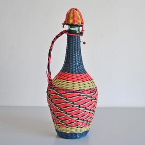 Op de afbeelding: Een vintage decoratieve fles met een geweven ontwerp. De fles heeft een blauwe basis, een rood en geel geweven middengedeelte en een geweven deksel. Een geweven handvat is aan de zijkant bevestigd. Een blikvanger voor interieurdecoratie.