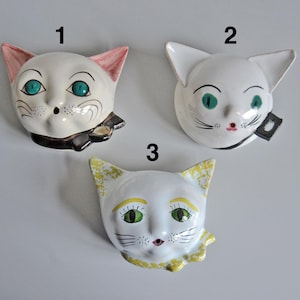 May include: Three ceramic cat head wall hangings. Each cat head is white with green eyes and black whiskers. Cat 1 has pink ears and a black bow. Cat 2 has a black collar. Cat 3 has yellow accents.