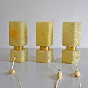 May include: Three vintage table lamps with a textured, yellow-gold finish. Each lamp has a rectangular base and a matching, slightly taller shade. The lamps are connected to a power source with a white cord and a switch.